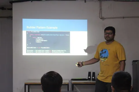 A man presents a slide titled Builder Pattern Example showing PHP code for a PizzaBuilder class to an audience.