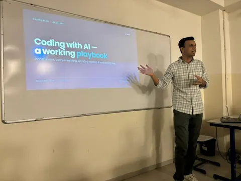 A man presents beside a slide titled “Coding with AI — a working playbook” at a community meetup.