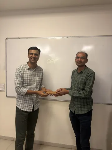 Two men smile while exchanging a rectangular plaque in front of a classroom whiteboard.